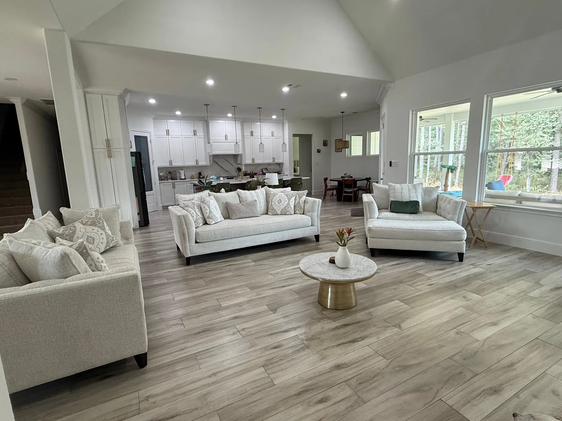 Great room under a vaulted ceiling with two pale sofas and a chaise, cushions plumped and pillows straightened, the tile floor clear through to the kitchen beyond